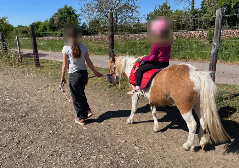 Ein Mädchen mit rosa Helm reitet ein Schecken-Pony, eine junge Frau führt es den Weg entlang
